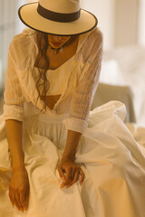 Elegant fashionable woman wearing summer white dress, straw hat, posing in stylish boho interior.