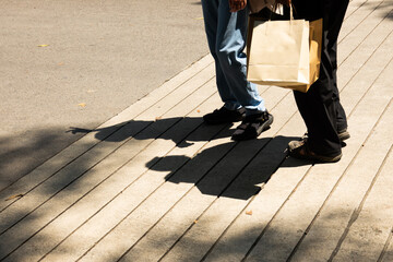 Two people walking on a sidewalk with one of them holding a brown paper bag. The scene is sunny and...