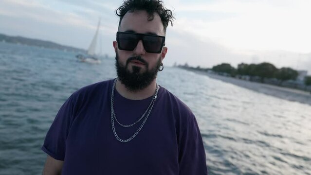 Dolly shot of a fashion-forward, bearded man in sunglasses and a chain standing by a river, gazing to the camera adds a bold touch to the serene setting.