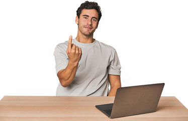 Hispanic young man working on laptop pointing with finger at you as if inviting come closer.