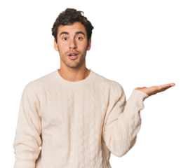 Young Hispanic man in studio impressed holding copy space on palm.
