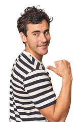 Young Hispanic man in studio points with thumb finger away, laughing and carefree.