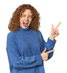 Young Caucasian redhead woman pointing with forefingers to a copy space, expressing excitement and...