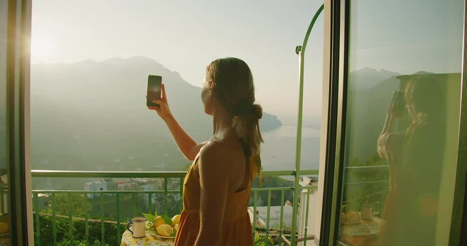 A woman in a yellow dress takes a photo with her phone from a balcony overlooking the Amalfi coast at sunrise.