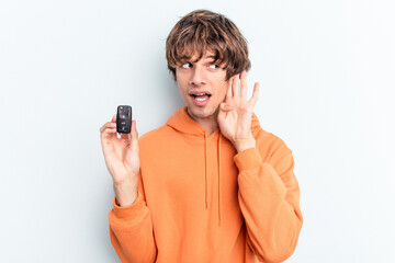 Young caucasian man holding car keys isolated on blue background trying to listening a gossip.