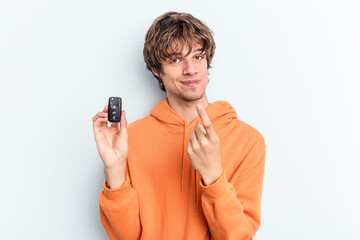 Young caucasian man holding car keys isolated on blue background pointing with finger at you as if inviting come closer. - obrazy, fototapety, plakaty