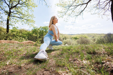 The girl is doing a stretching squat on one leg. Beautiful blonde Caucasian woman in blue tight tracksuit. Blonde girl at an outdoor training session