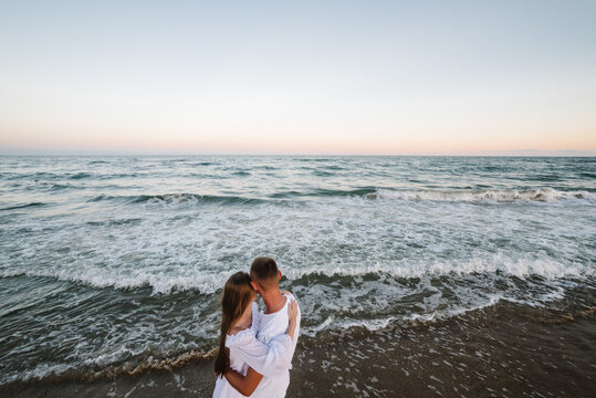Love. Couple hugging on seashore in sea. Male hugs female standing on water with big waves ocean and enjoying a summer day. Man embraces woman walking on beach sand sea. Top back view.