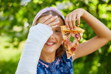 Smiling Young Girl With Cast Enjoying Pizza Outdoors on a Sunny Day