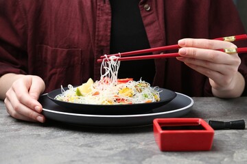 Stir-fry. Woman with chopsticks eating tasty rice noodles with meat and vegetables at grey textured table, closeup