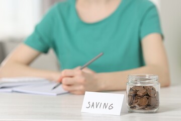 Financial savings. Woman making notes at white wooden table indoors, focus on glass jar with coins