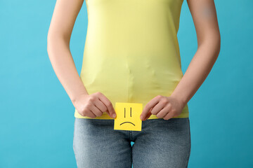 Cystitis. Woman holding sticky note with drawn sad face on light blue background, closeup