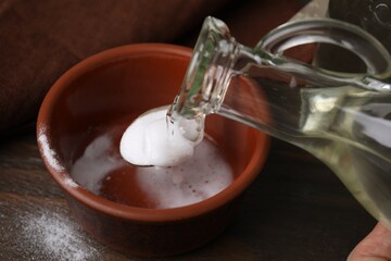 Fototapeta na wymiar Pouring vinegar into spoon with baking soda over bowl at wooden table, closeup