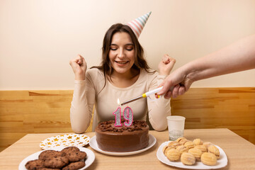 A girl sits in front of a table with a festive cake, in which a candle is lit in the form of the number 19. The concept of a birthday celebration.