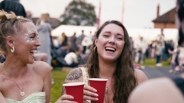 Three female friends wearing glitter and doing cheers with drinks talking and having fun at outdoor summer music festival - shot in slow motion 