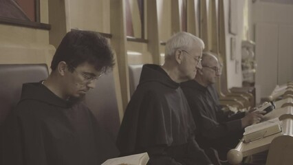 Monks reading Bible together while sitting in church side view medium shot. Christian monks praying liturgy with psalmbook. Liturgy of the Hours concept
