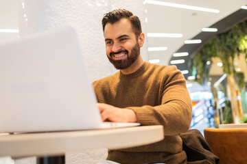 A cheerful man works on his laptop in a modern cafe setting, his smile suggesting productivity and satisfaction with his work