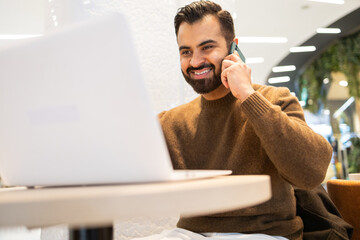 Engaged and personable, a man conducts a phone conversation with a warm smile, seated with his laptop in a contemporary cafe setting