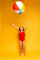 Happy little girl wearing red swimsuit and sunglasses playing with ball on yellow background. Summer vacation and childhood concept. Active lifestyle