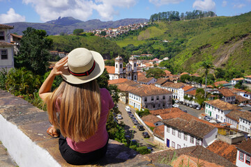 Tourist girl with hat sitting on wall looking at panoramic view of the historic city of Ouro Preto,...