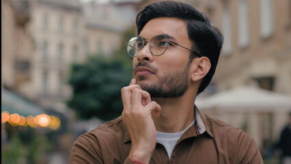 Close up thoughtful confused puzzled Indian Arabian ethnic male man student businessman guy deeply thoughts pondering thinking business solution idea problem think outdoors city cafe restaurant street