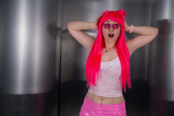 woman in glasses, dressed like a doll. Young beautiful sexy woman in camisole and pink skirt on silver background. Red hair girl wears pink wig with fringe. Showing different emotions