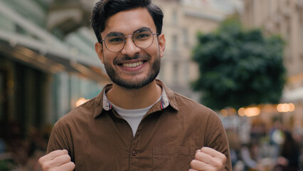 Excited Indian Arabian ethnic male man 20s guy bearded businessman happy student celebrating achievement victory clenching fists gesture good news winning lottery looking at camera outside city street