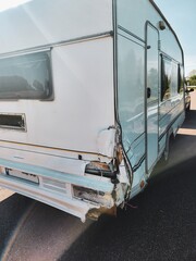Vintage white travel trailer with a unique retro design and a dent on the back corner, parked by a tranquil lake surrounded by lush green trees, creating a serene setting for a peaceful retreat.