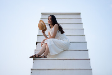 A smiling woman dressed in white sits casually on a staircase, holding a straw hat, exuding a...