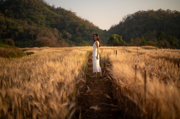 A young woman in a white dress and straw hat smiles while standing amidst a field of golden wheat,...