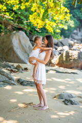 Young woman mother with a little daughter in white dresses on seashore in the shade of trees and palms