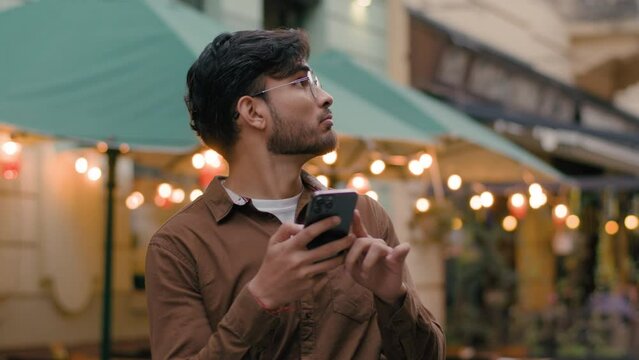 Nervous lonely Indian Arabian ethnic male businessman guy looking around feeling lost. Man holding smartphone use mobile electronic map geolocation app trying find location outside city evening street