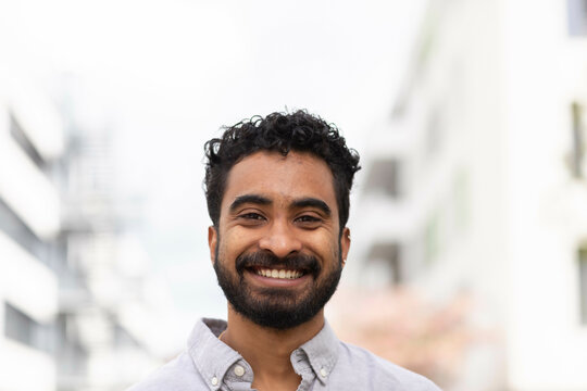 A cheerful man with a beard and curly hair is smiling at the camera in front of a blurred urban background.