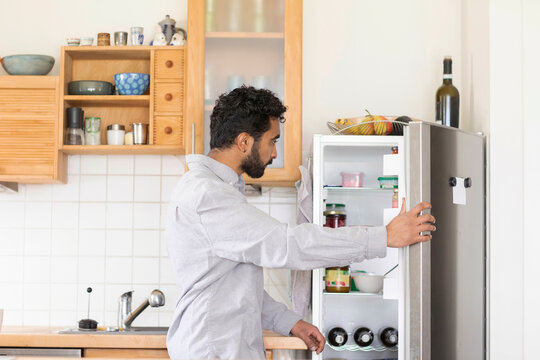 A man is opening a refrigerator full of various food items in a well-lit, modern kitchen.