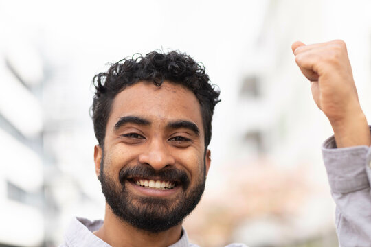 A smiling man with curly hair is raising his fist in a gesture of success or celebration against a blurred urban background.