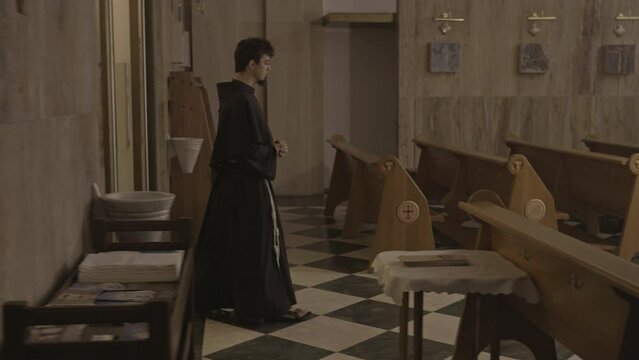 Young caucasian christian monk walking in church side view tracking shot. Clergyman wearing brown robe navigating through wooden pews rows aisle in cathedral