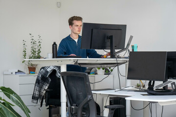 A man stands at an adjustable desk in a modern office, working on a computer with dual monitors.