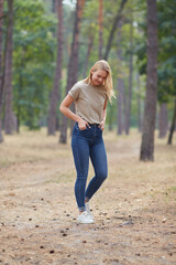 Blue-eyed blonde in a beige T-shirt walks in a pine forest. Portrait of a joyful young woman enjoying in autumn park.