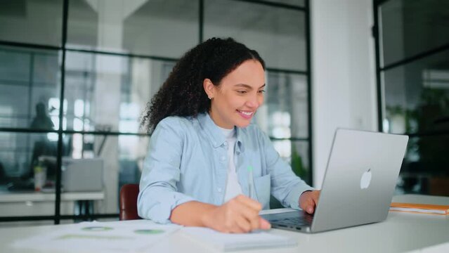 Motivated positive gorgeous brazilian or hispanic curly woman in a pastel blue shirt, employee of a company, sits in a modern office at work desk, working on a laptop, takes notes in notebook, smiles