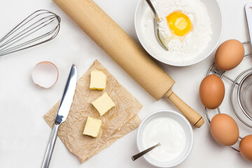 Brown eggs on metal stand. Butter and knife on paper. Egg yolk with flour and spoon in bowl. Milk...