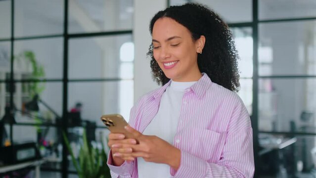 Positive brazilian or latino curly haired business woman, corporate manager, designer, walking in modern office with smart phone in hands, using gadget, looks at it, typing text, smiles