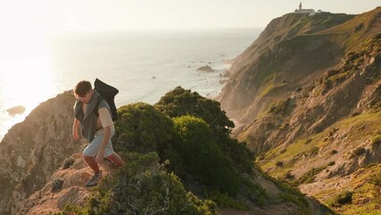 A young hiker with a backpack climbs a steep hill beside the ocean during sunset, embracing the adventure and scenic view.