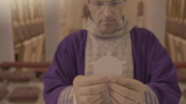 Priest lifting sacramental bread, celebrating Eucharist in church close up handheld shot. Clergyman in purple robe holding communion host in hands closeup