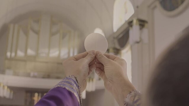 Christian priest lifting sacramental bread selective focus view over shoulder. Catholic clergyman holding communion wafer in hands during Eucharist celebration ceremony