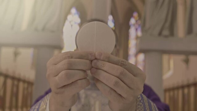 Chaplain lifting communion bread from golden plate on altar close up. Christian priest holding sacramental host in hands during Holy Communion ceremony ritual