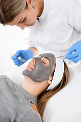 Doctor in a blue rubber gloves makes a face mask for a woman with the help of cosmetics tools - 791698687