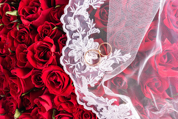 Background image for wedding design or mockup. Two gold wedding rings on a white bridal veil. Top view. red background roses bouquet. A copy space