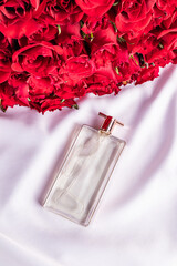 A transparent chic perfume bottle with a delicate fragrance lies on a white satin background. red roses bouquet. Top view. Presentation . Blank lay.