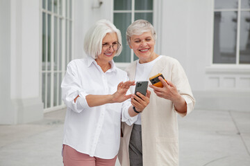 Two joyful senior women standing outside of building, smiling exchange contact details using smartphones, wearing stylish casual attire, enjoying warm sunny day together. Happy mature female model