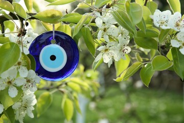 Evil eye amulet hanging on blossoming spring tree outdoors, closeup. Space for text